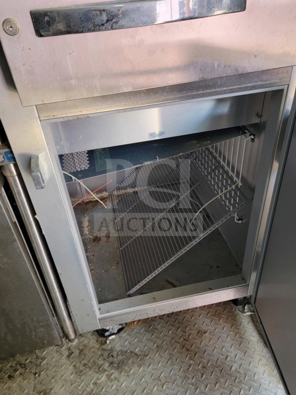 Open stainless steel refrigerator with wire shelves inside a mobile kitchen trailer, showing moderate wear and dirt.