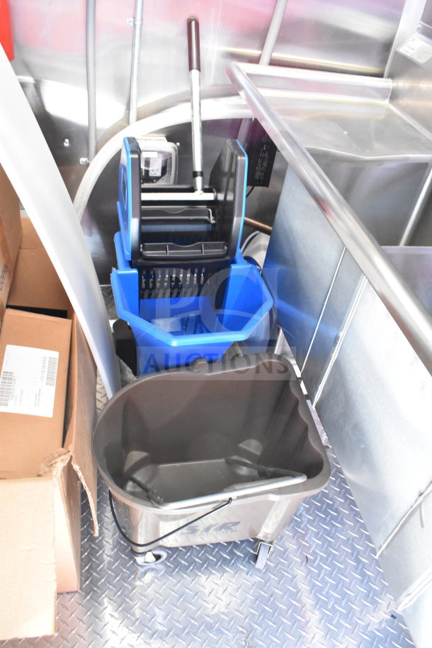 2022 Rock Solid Cargo trailer interior with cleaning supplies, including a blue mop bucket and brown waste bin on metal flooring.