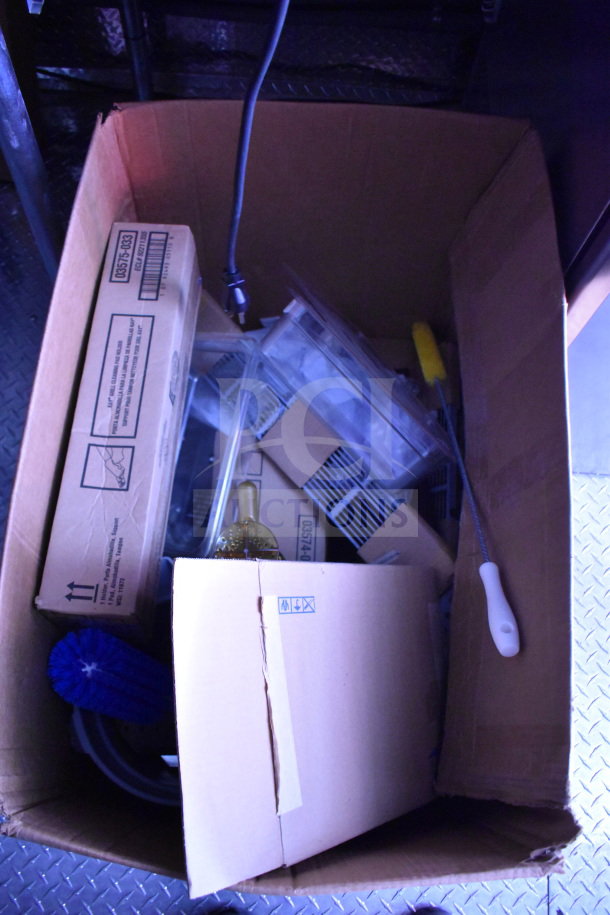 Cardboard box with assorted food trailer accessories, including brush and labeled packaging, inside Rock Solid Cargo trailer.