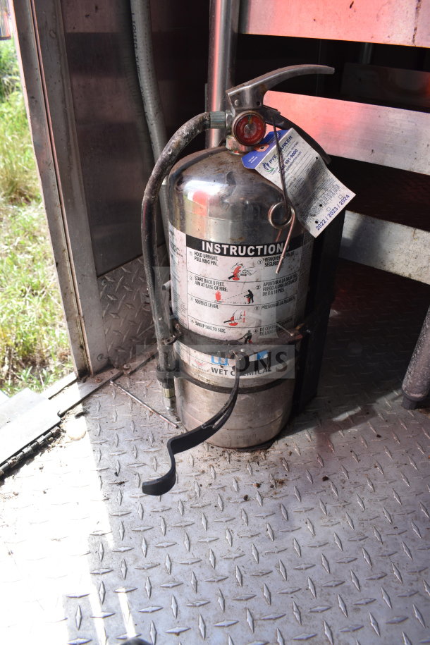 Fire extinguisher inside a 2021 Titanium Cargo Trailer with instruction label and inspection tag visible, set on metal flooring.