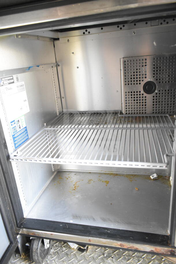 Interior of a True refrigeration unit in the 2021 Diamond Cargo 8.5'x24' food trailer. Features include a wire shelf and visible grease stains, suggesting prior use. Labels indicate model and maintenance instructions.