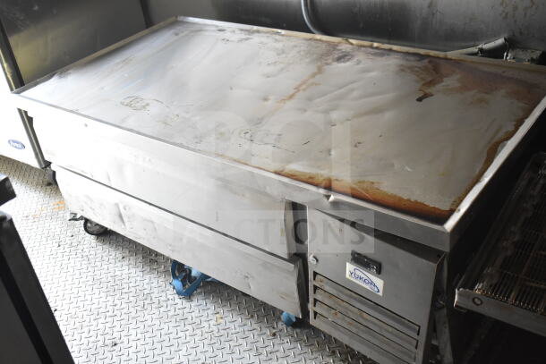 Stainless steel commercial griddle on wheels, showing signs of wear. Visible "Yukon" label on lower compartment.