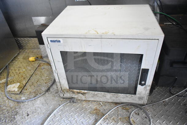 White metal cabinet on diamond plate floor with visible dirt and electrical cords. Marked "martin."