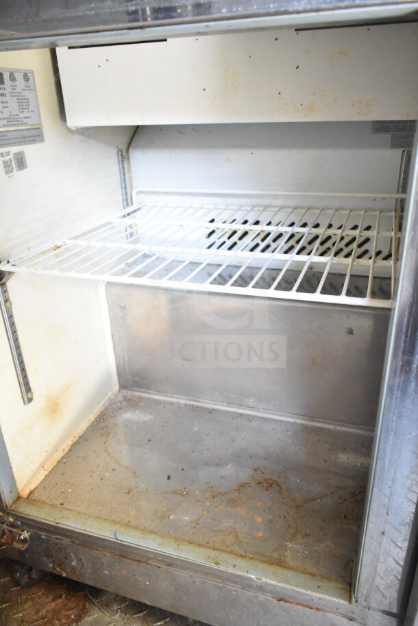 Interior of a used food trailer refrigerator, shows some rust and wear. White wire rack shelving. Labels visible on the side.