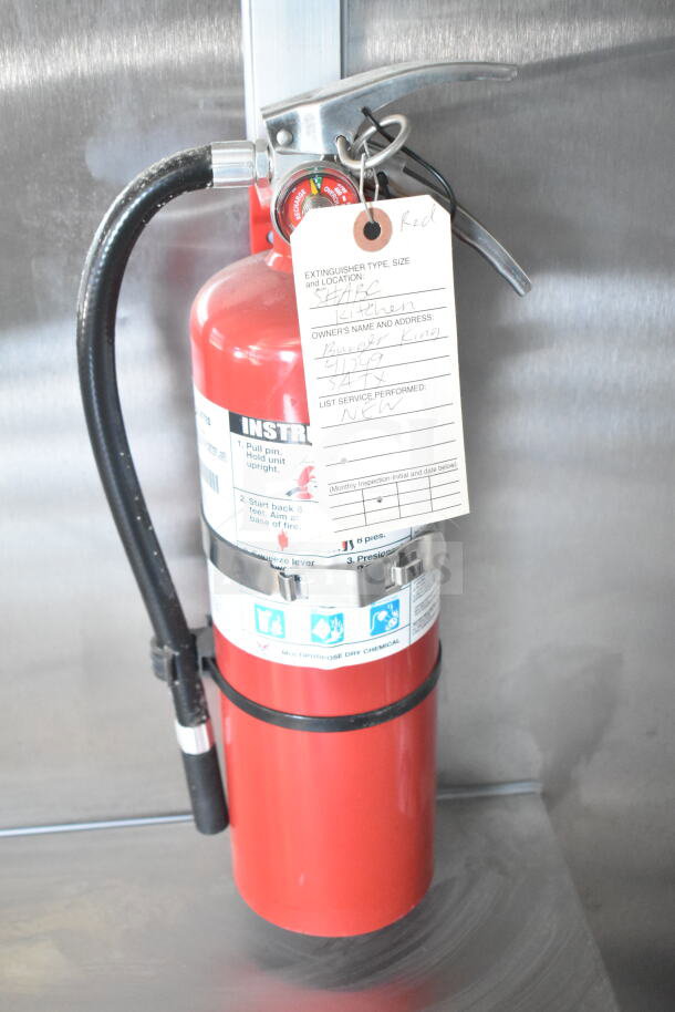 Red fire extinguisher with attached service tag listing type as ABC and location as kitchen; secured in metal bracket.