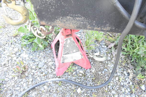 Red jack stand with label beneath a trailer hitch, chain, and hook on gravel with weeds.