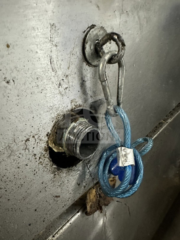 Stainless steel surface close-up with a gas connection fitting and attached blue cable, showing wear and tear.