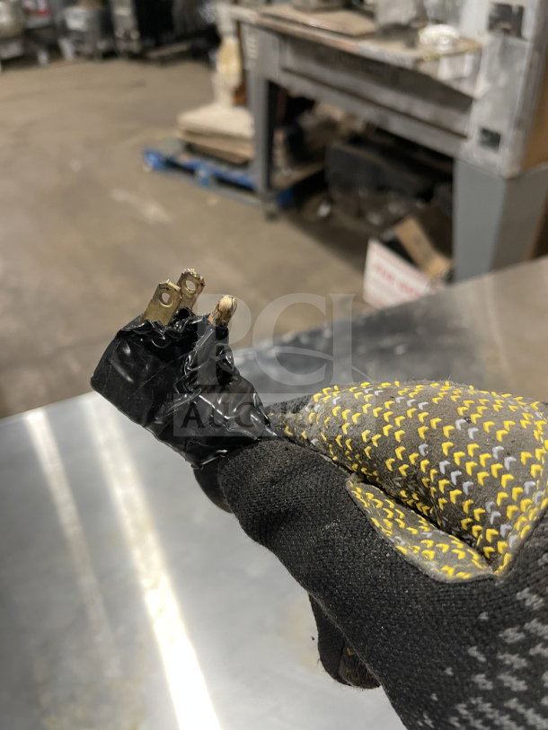 A close-up of a damaged electrical plug wrapped in black tape, held by a gloved hand in an industrial setting.