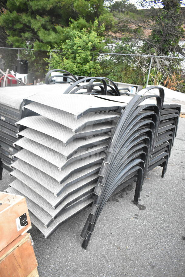 Stacked 22 outdoor chaise lounge chairs with metal frames and woven seats, in used condition, on a paved surface.