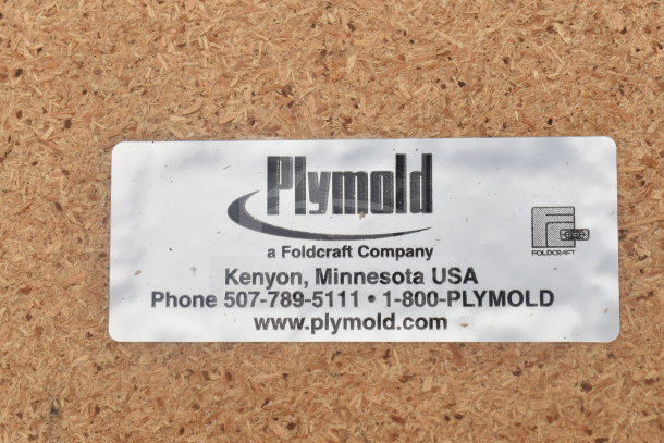 Plymold label on wood surface. Text includes contact info: Kenyon, Minnesota, USA, phone numbers, and website www.plymold.com.
