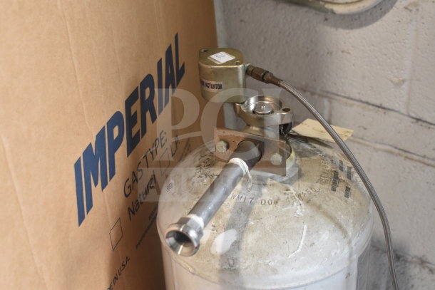 Fire suppression tank with actuator, connected hoses, and visible labeling. Positioned adjacent to an "Imperial" box.