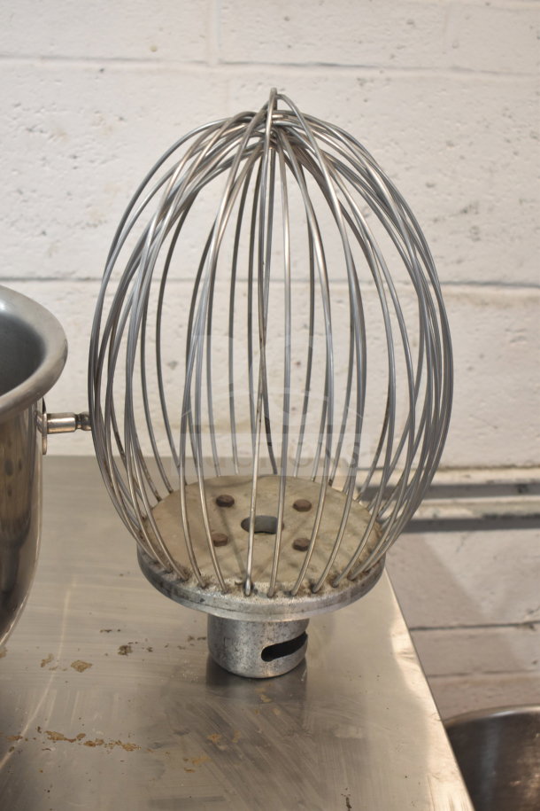 Stainless steel whisk attachment for mixer on metal surface, showing durable construction and well-used condition.