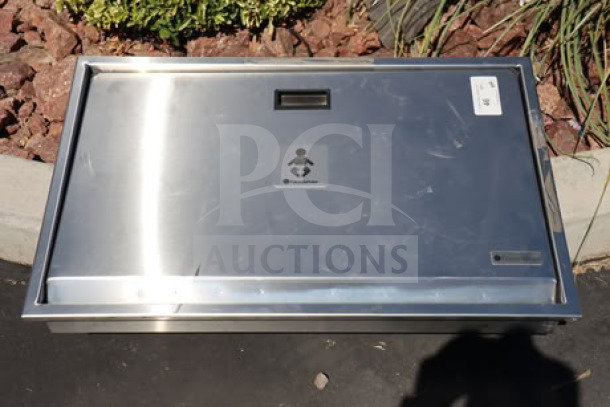 Stainless steel baby changing station, wall-mounted, with child icon label, excellent condition, slight surface marks visible.