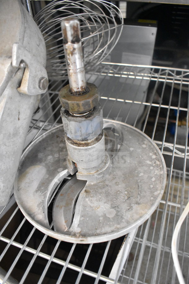 Metal Pelican head with S blade and whisk attachment on wire shelf, slightly worn, ideal for food processing tasks.