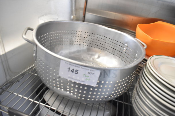 Aluminum colander with handles, labeled "Tier Lot 145," stacked with plates and baking pans on a metal shelf.