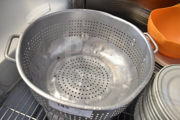Metal colander with handles, stack of ceramic plates, and orange mixing bowl on a shelf; used condition.