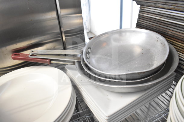 Alt text: Tiered stack of mixed kitchenware including metal colanders, frying pans with red handles, white plates, and square baking pans on wire rack.