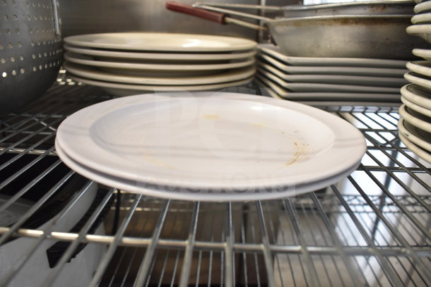 Stack of white plates and metal colander on wire shelf, includes baking pans. Plates show slight wear, ideal for bulk kitchen use.