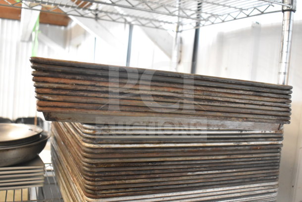 A stack of metal baking pans in used condition on a wire shelf, part of a mixed lot with plates and a colander.