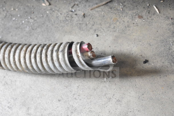 Coiled metallic electrical conduit with exposed wires on a dusty concrete floor. Visible signs of wear.