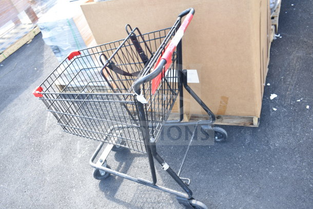 Black metal shopping cart with red handle, in good condition, features four wheels and child seat, standing on asphalt.