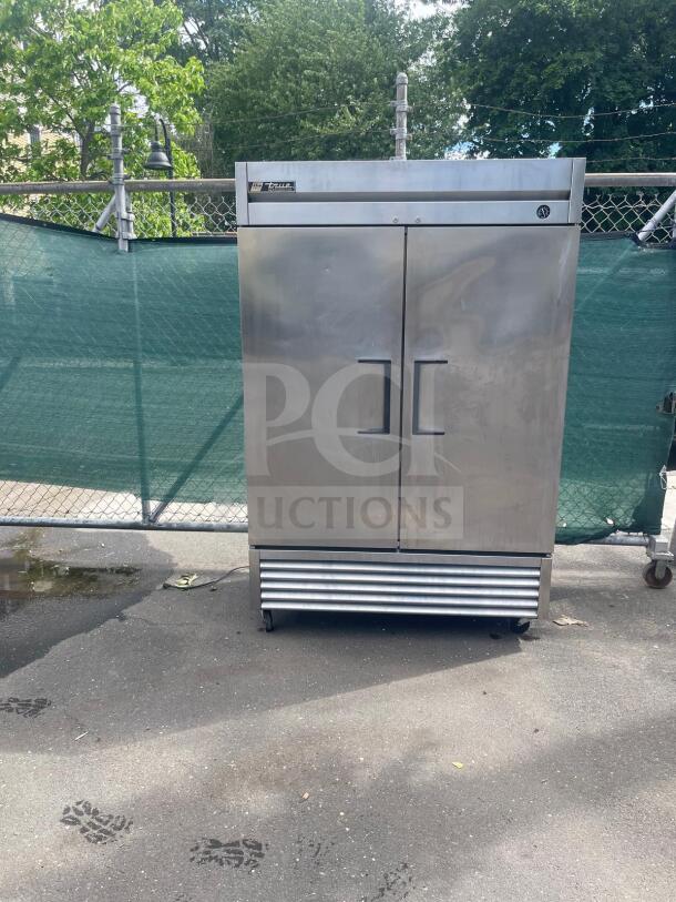 True double-door commercial refrigerator, stainless steel, on wheels, visible wear. Brand label on top left corner.