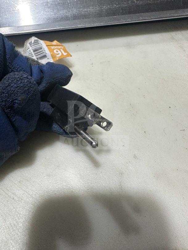 A gloved hand holding a metal prong next to a surface; a partially visible orange tag with barcodes is in the background.