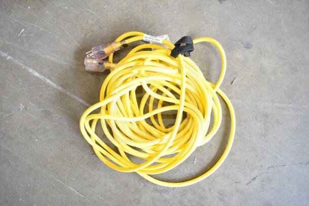 Yellow extension cord coiled on a concrete floor, featuring dual three-prong transparent plugs and a black cable tie.