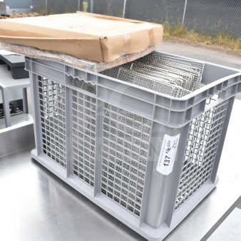 Brand new stainless steel mesh tray racks in a gray plastic bin; lot label suggests 50 units, scratch and dent condition.