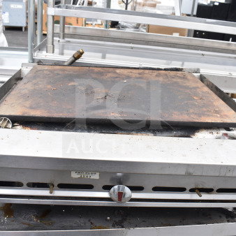 Commercial countertop gas-powered flat top griddle with metal casing. Shows signs of use; includes temperature control knob and vent slots. Serial tag visible.