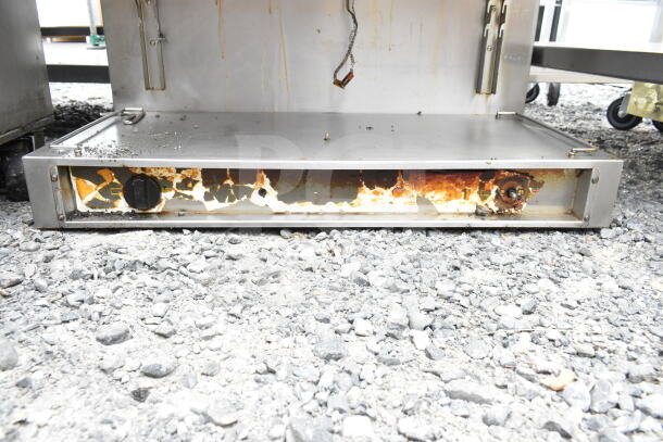 Commercial cheese melter with visible rust and wear on lower panel, located outdoors on gravel.