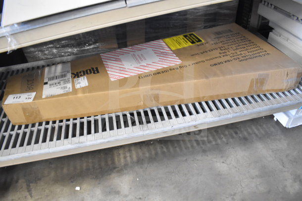 Cardboard box labeled "Rock Jam Guitar" on a metal shelf, auction lot 113. Box has labels and shipping tags attached. Condition appears unused.