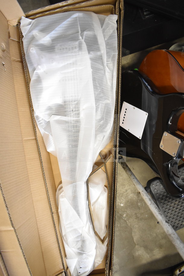 Rock Jam guitar wrapped in white plastic inside a cardboard box, partially visible with glossy black and brown finishes.