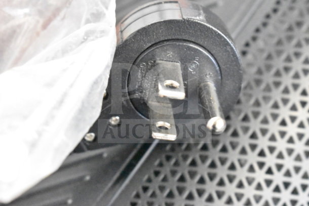Close-up of an electrical plug for JBL PartyBox 110 speaker, resting on a textured surface, showing prongs and markings.