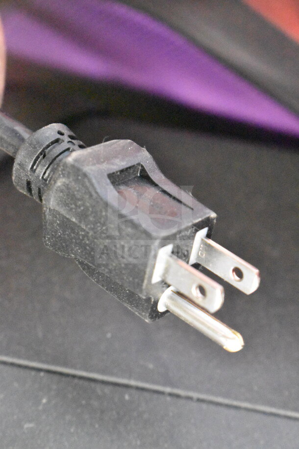 Power plug close-up, black plastic, two prongs visible on the connector, against blurred dark background.