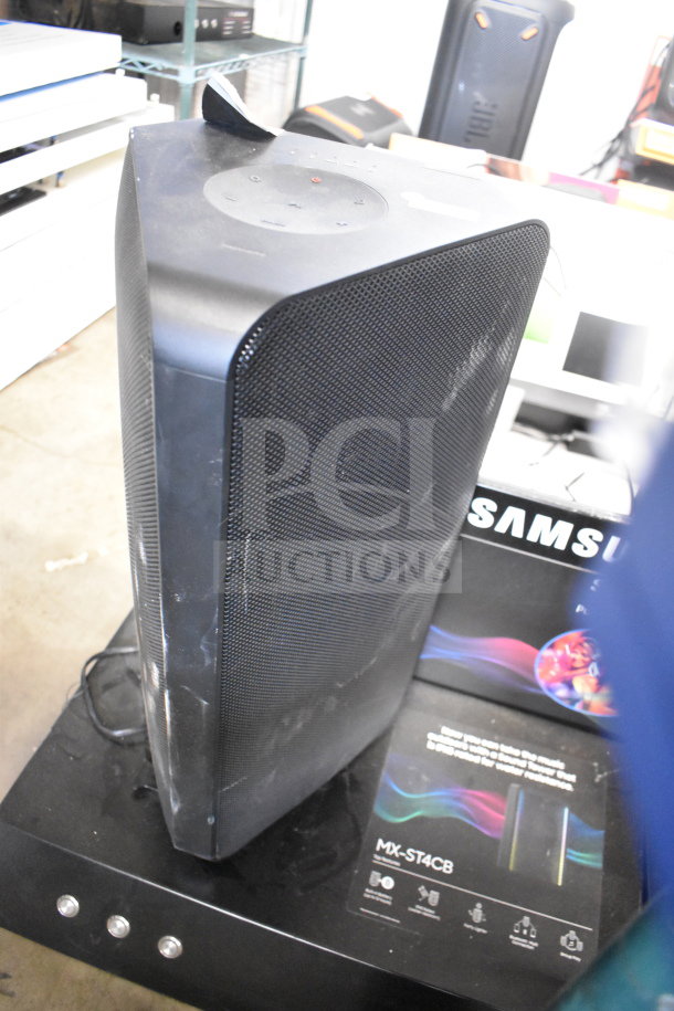 Samsung MX-ST4CB speaker on store display, black finish, visible wear, on top of a promotional poster, emphasizing sound quality.