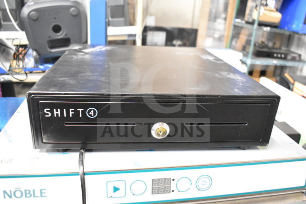 Shift4 cash drawer, black metal with visible lock and slots, in used condition on top of a Noble box.