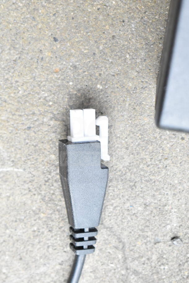 Close-up of a black electrical cord with a white connector, part of a lot of cords and battery charger in a clear poly bin.
