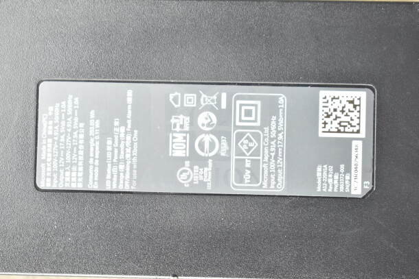 Clear poly bin containing various cords and a battery charger. Label shows input/output specifications for Microsoft Japan Co., Ltd.