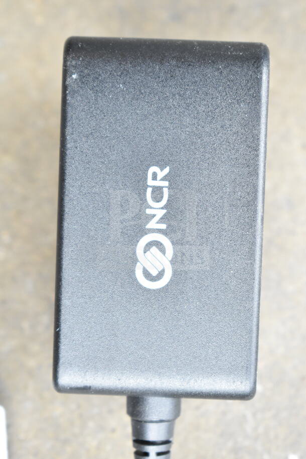 Clear poly bin containing various cords and a battery charger. Charger with NCR logo. Ideal for tech enthusiasts.