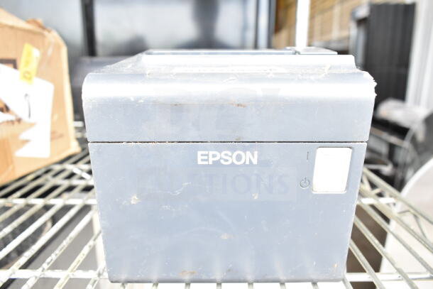 Epson receipt printer on metal shelving, used condition, power button visible. Box in background with partial label.