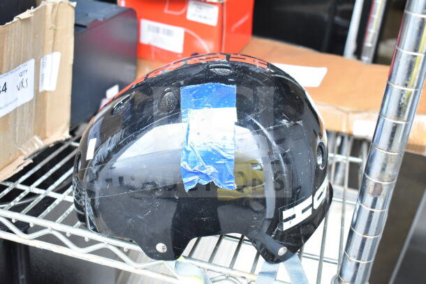 Black OnlyNY helmet with visible scratches and blue tape, resting on a metal shelf amidst boxes.