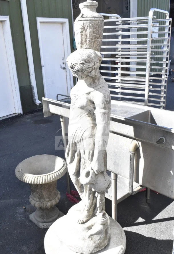 Stone statue depicting a classical figure holding a jug, detailed engraving on base, weathered condition, approximately 4 feet tall.
