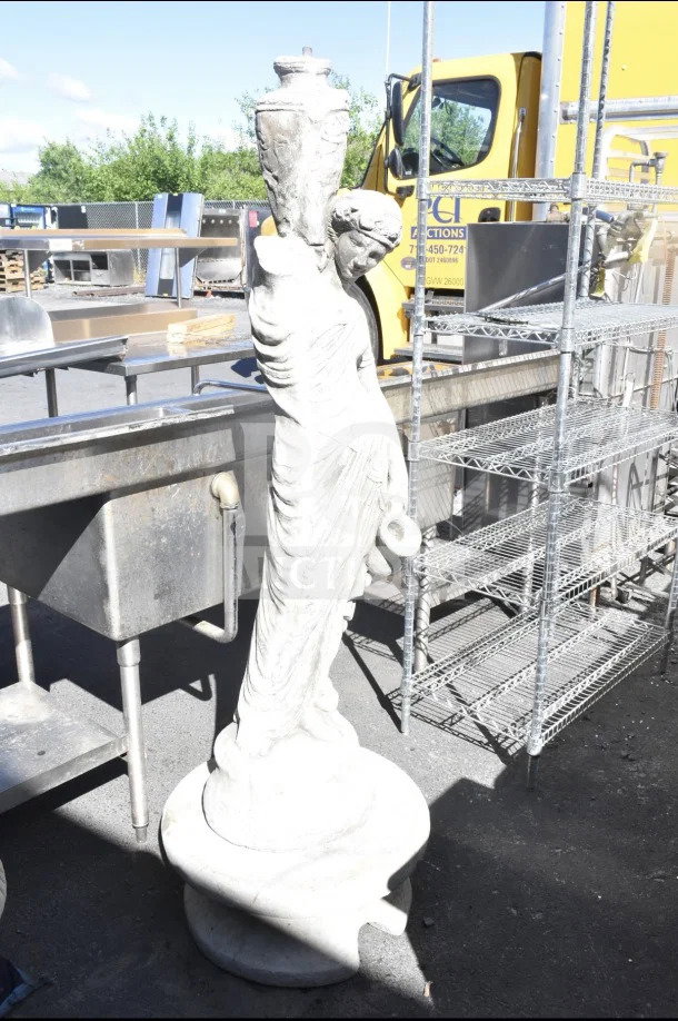 Stone statue depicting a draped figure holding a jug, standing on a round base. Weathered appearance, outdoor setting.