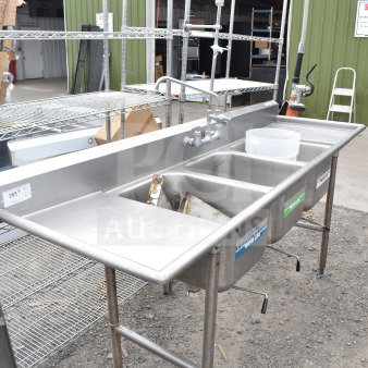 Commercial 3 bay sink with dual drain boards, faucet, and spray nozzle. Stainless steel, durable, used condition.