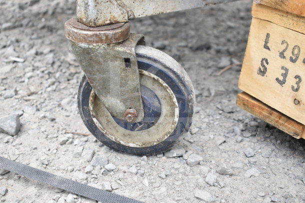 Commercial pan transport rack caster, worn condition, on a gravel surface. Industrial metal and rubber wheel detail.