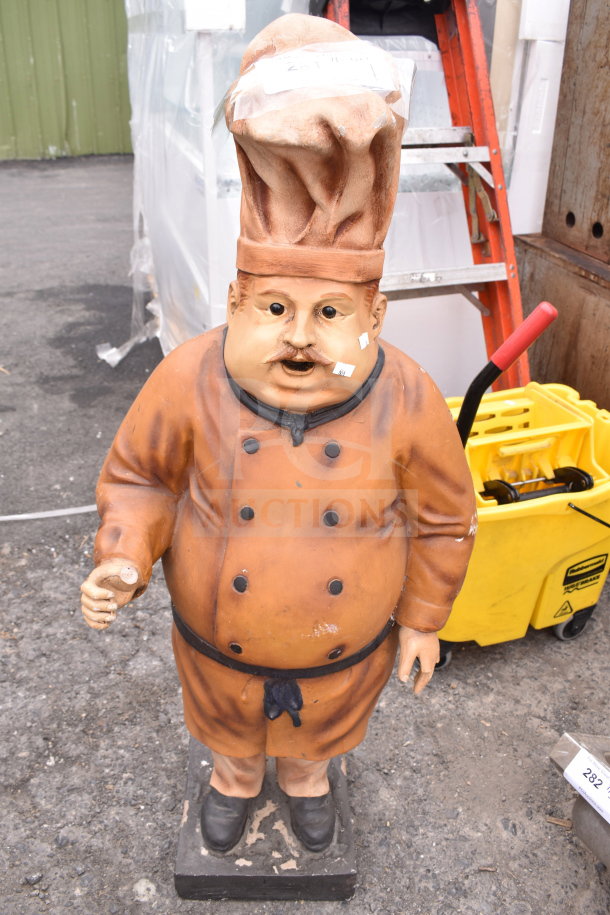 Lifesize chef statue in orange uniform and hat, holding a spoon. Slight wear visible. Unique decor item for kitchens.