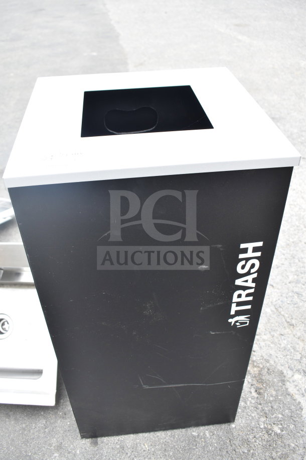 Black trash bin with a white top and "TRASH" label on the side. Shows minor surface scuffs.
