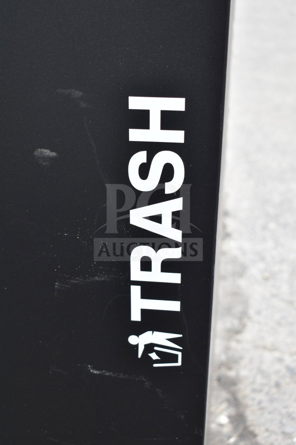 Black trash bin labeled "TRASH" in white text, visible signs of wear. Suitable for outdoor or commercial use.