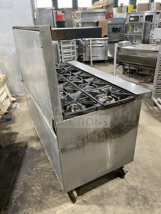 Bakers Pride stainless steel 10-burner gas range with double oven, model 955394604036, on casters. Includes backsplash and overhead salamander shelf. Visible wear.